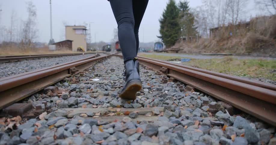 Woman with jeans and black boots walks along rusty railway tracks on a cloudy day