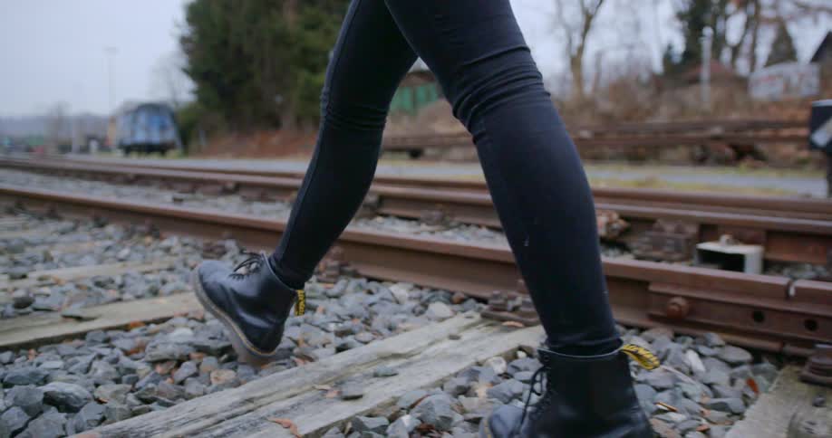 Woman with jeans and black boots walks along railway tracks on a cloudy day
