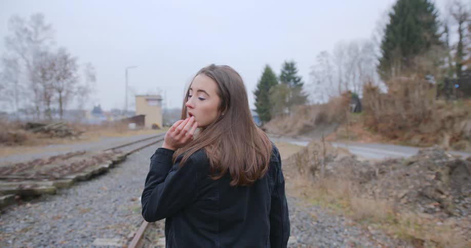 Woman with brown hair and a denim jacket walks along railway tracks on a cloudy day