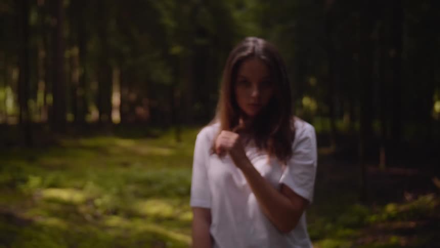 Girl with brown hair and a white shirt walking in the woods turns and looks at the camera