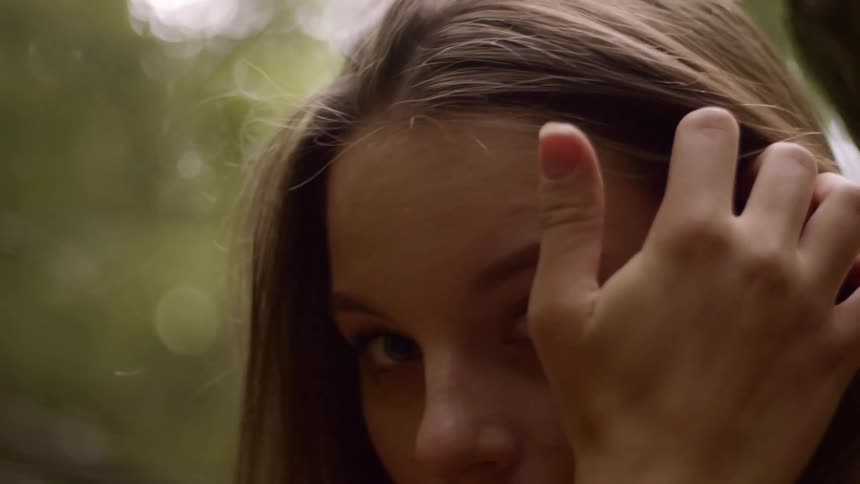 Teenage girl with brown hair and a white shirt touches her hair and looks at the camera in the forest