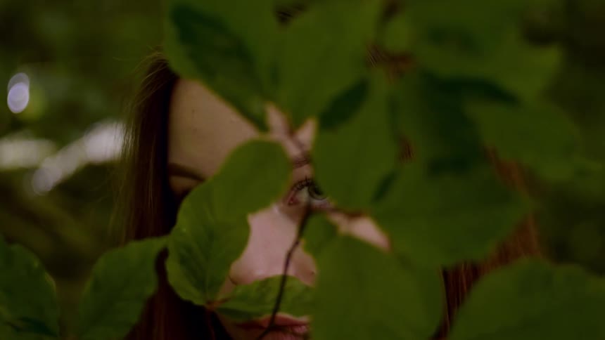 Teenage girl with brown hair looks at the camera from behind leafy branches in the forest
