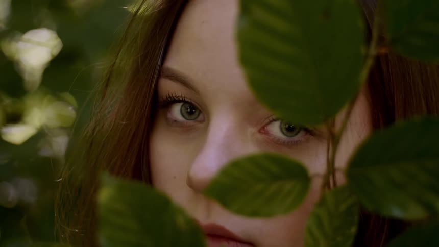 A woman poses in a garden and obscures her face with leaves on a plant