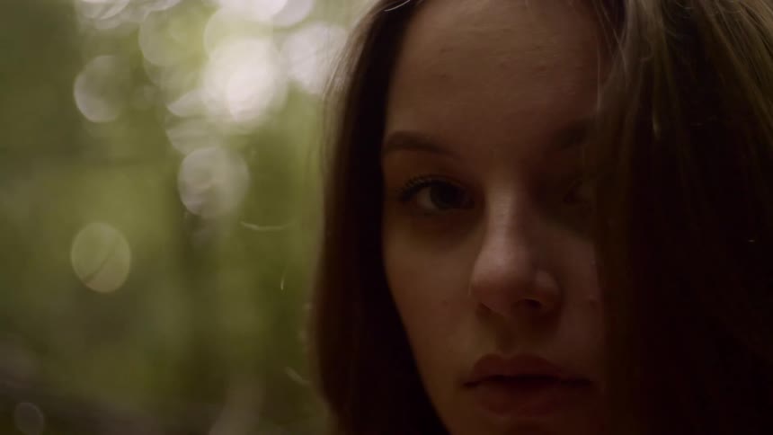 Teenage girl with straight and brown hair stands out against a blurry background and looks at the camera in the forest