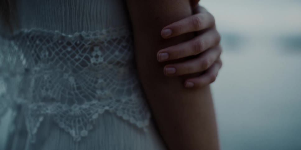 A woman in a white dress slowly runs her hand down her arm while standing outdoors