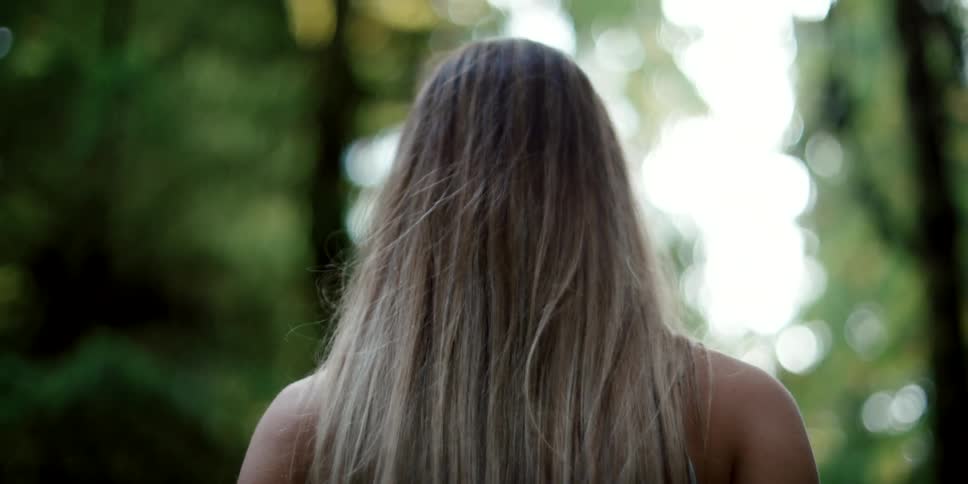 Woman with blonde hair looks up at tall trees in the forest
