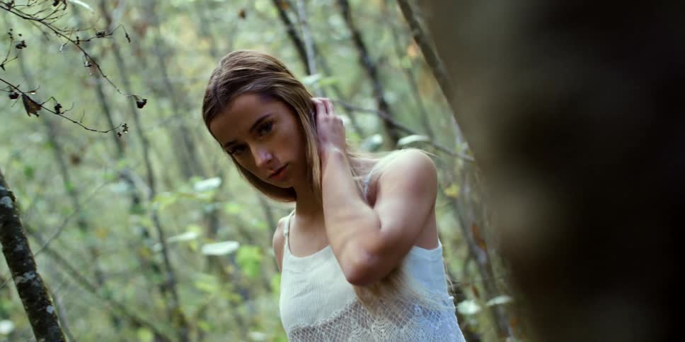 A young woman brushes her hair behind her ear while walking through a woodland and looking into the camera