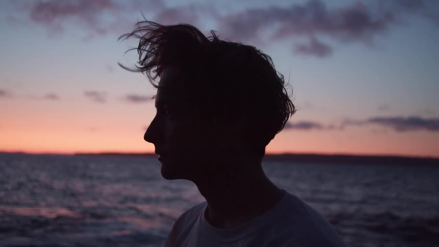 A young man looks at the ocean at sunset, his hair blowing in the wind.