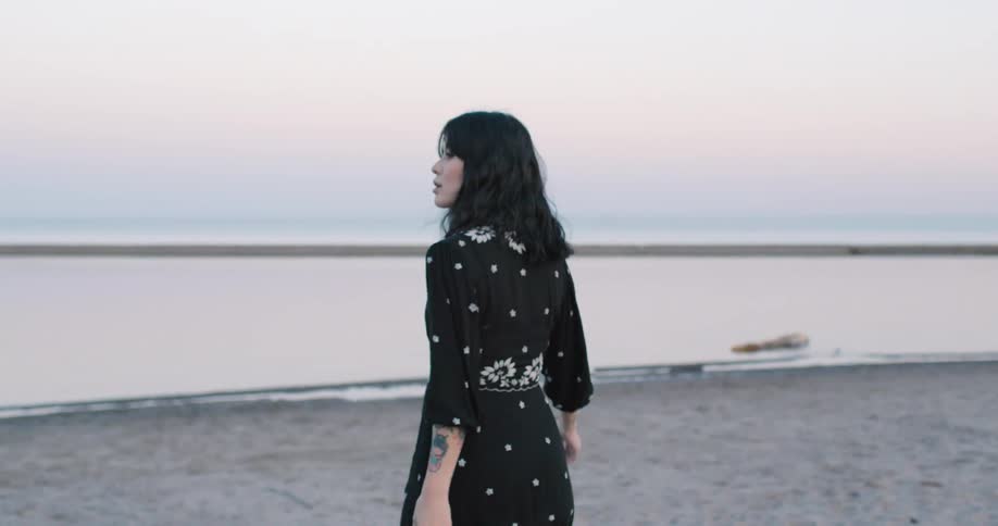 Asian woman with a black dress walks on towards the water on a beach at sunset