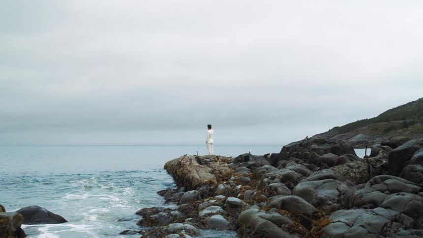 A person stands on the rocky shoreline looking out at the ocean