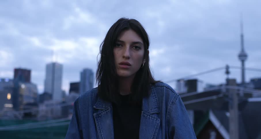 A white woman stands on a city rooftop looking into camera.