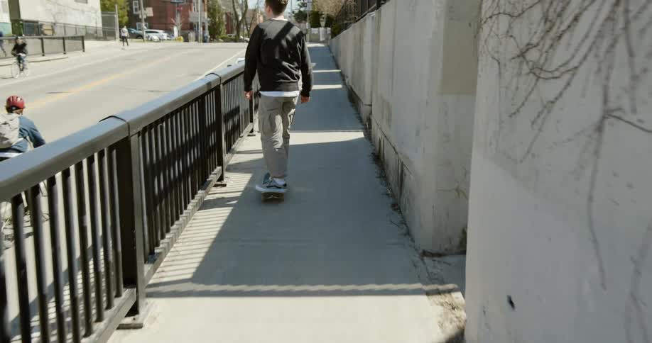 Teenager skateboards on the sidewalk along a road under bridge.