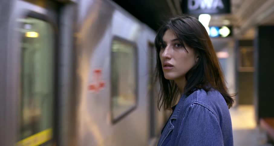 A young woman's hair blows in the wind of an approaching subway train entering the station as she waits on the platform