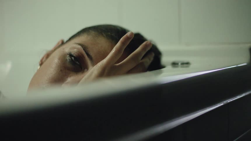 A young woman in the bathtub looking distressed, sinking under the water.