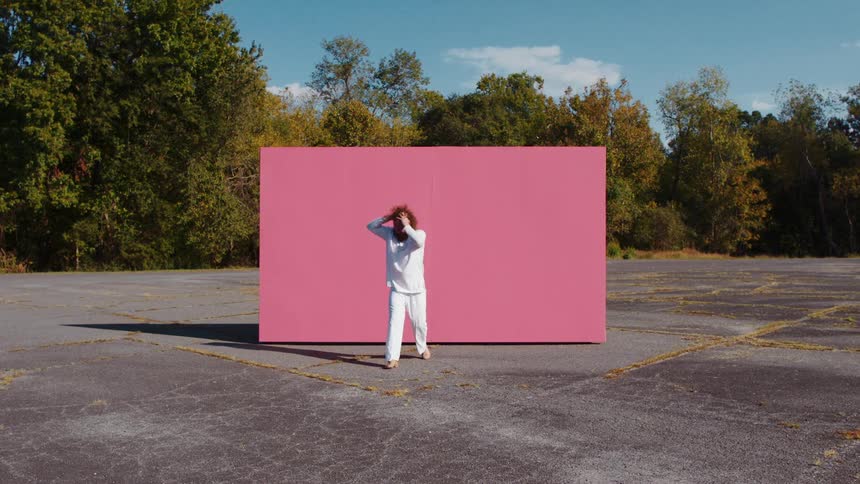A barefoot man in white clothes dances in a parking lot in front of a pink screen on a bright sunny day