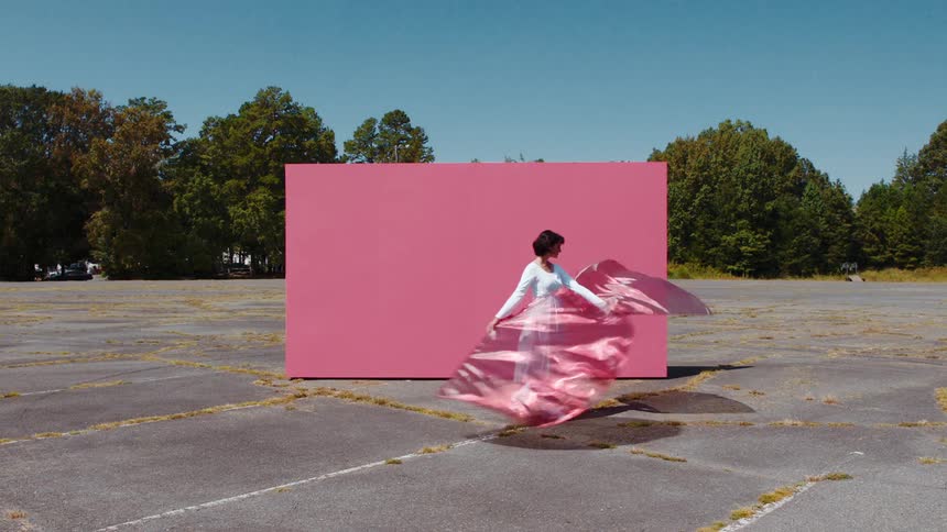 Woman dressed in white dances with pink translucent flags in front of a pink backdrop in a parking lot