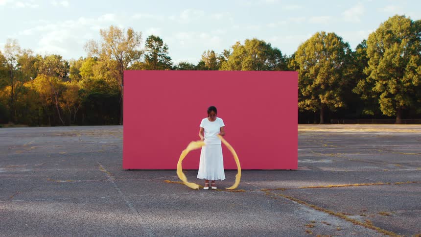 A woman in a white dress performs a ribbon dance in front of a red screen in a vacant parking lot