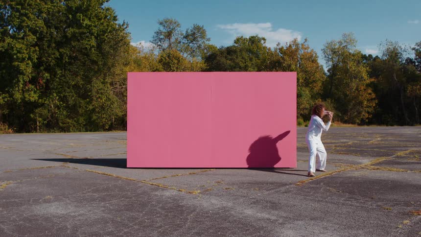 Bearded man in white linen shirt and pants dances barefoot in empty lot in front of blank pink sign