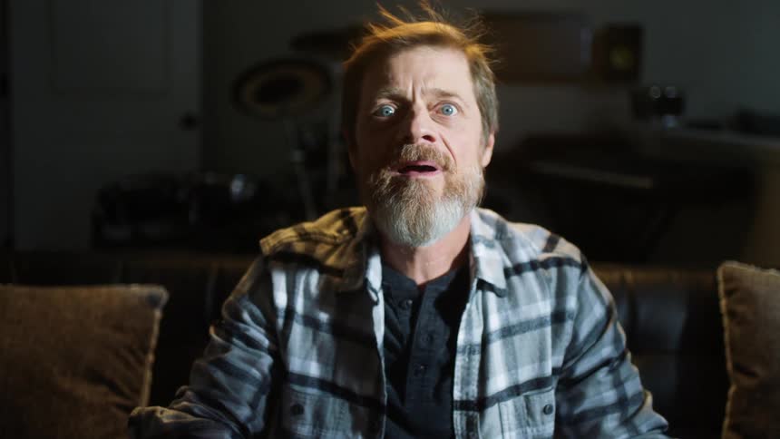 Man holds a remote while sitting on a sofa and looks surprised