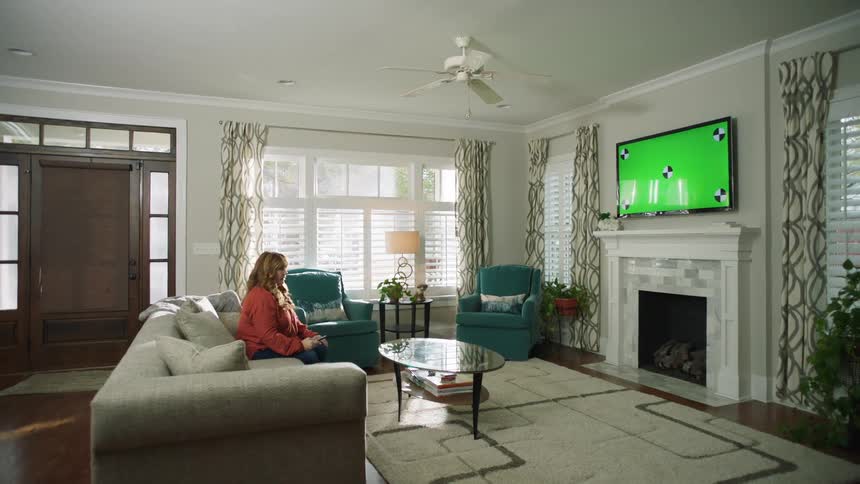 A woman sits on a couch holding a remote looking a television