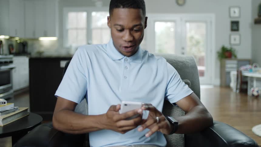 Happy black man with a blue polo shirt and beige pants sits in an black armchair picks up a smartphone from a black occasional table