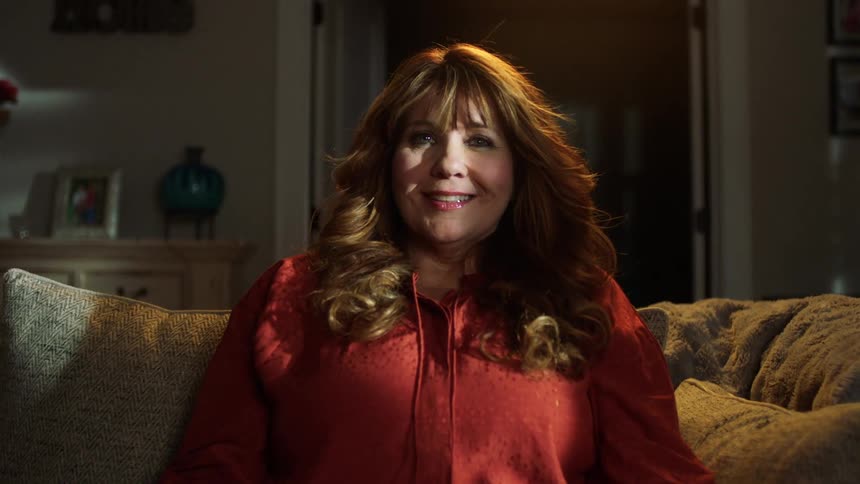 A woman sitting on her sofa smiling as her long hair and bangs are blown by a fan or wind machine