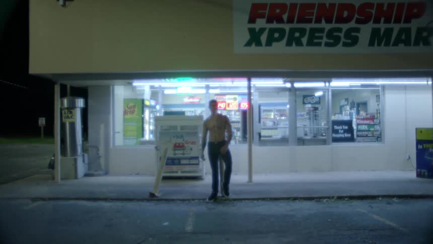 Barechested man stands outside convenience store near parking lot smoking