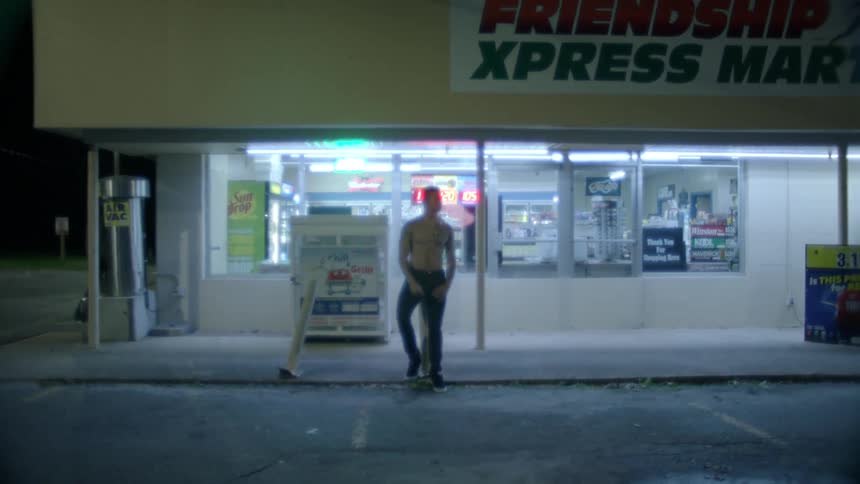 A white man stands in front of a convenience store smoking a cigarette at night