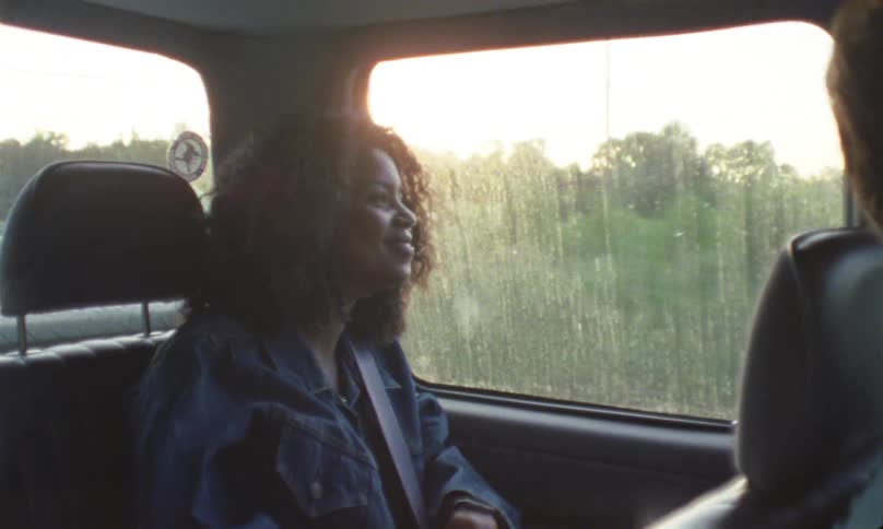 Black woman with curly hair sitting in a car looks out the window of a moving car and laughs as the sun goes down