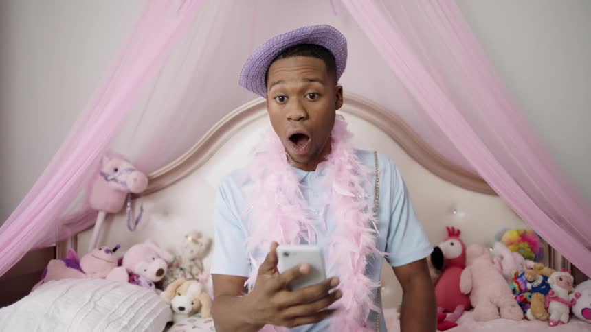 A young man dressed up in a hat and feather boa sits on a bed filled with stuffed animals and looks at his cell phone