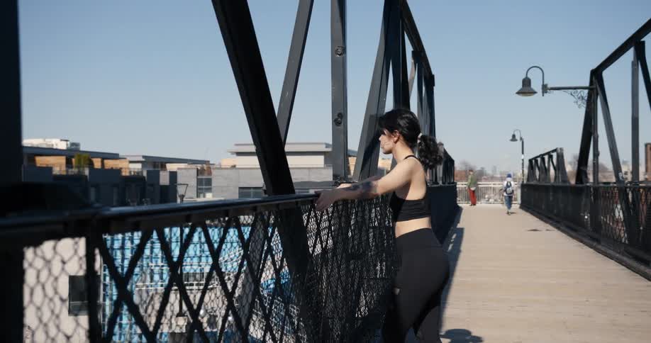 A female jogger stretches and catches her breath on a pedestrian bridge over train tracks.