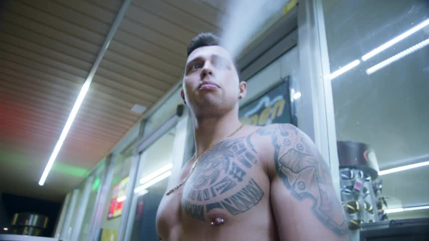 A shirtless man smokes a cigar in front of a convience store