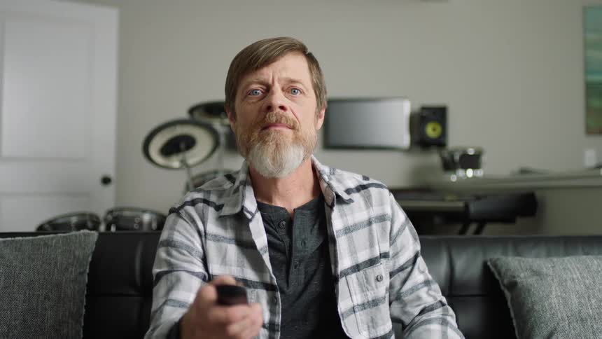 Man sits on a black sofa and switches on the television with a remote control