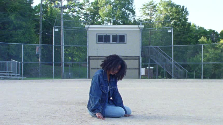 A woman sitting in the dirt of an empty sports field is drawing on the ground
