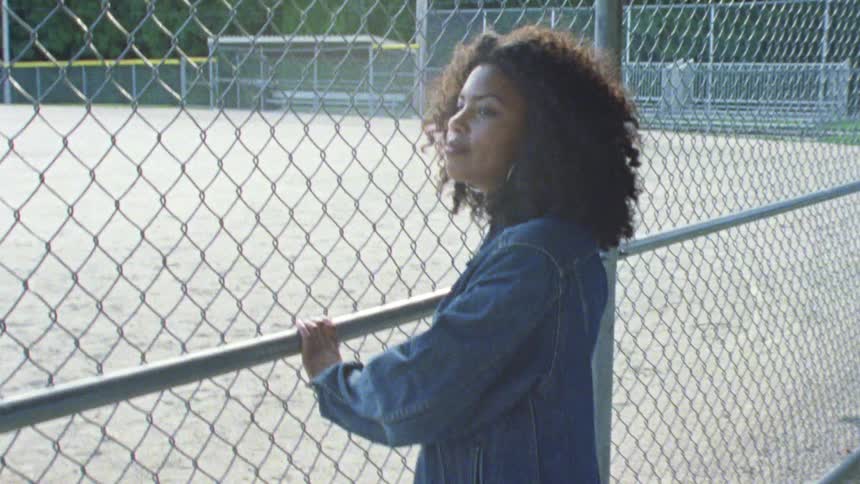 Young woman wearing denim jacket leaning on a wired fence in a baseball field at sunset