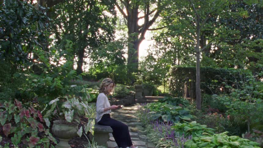 A woman sits outdoors on a stone bench in a large garden reading a book