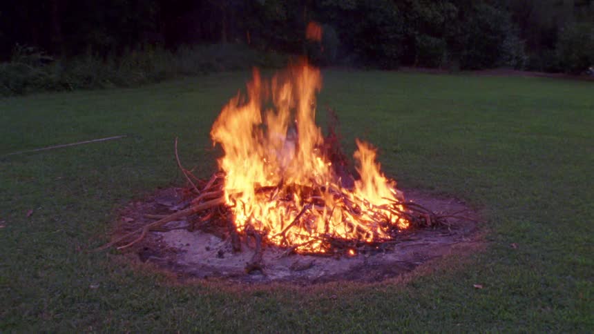 Fire burns outside on mowed lawn bordered by trees