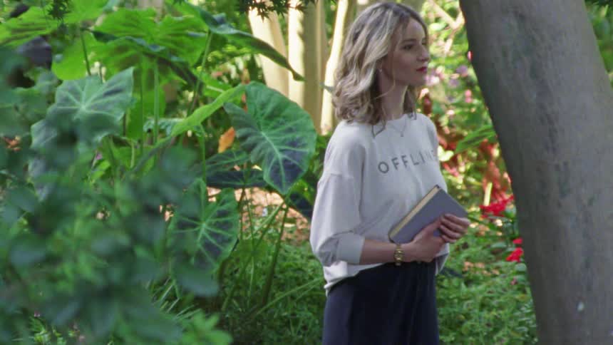 A woman stands in a garden holding a book and waiting for someone