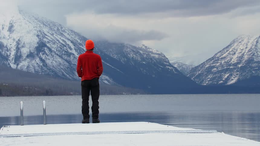 A young man walks to the edge of a dock and looks down at the water and then across to the mountain range