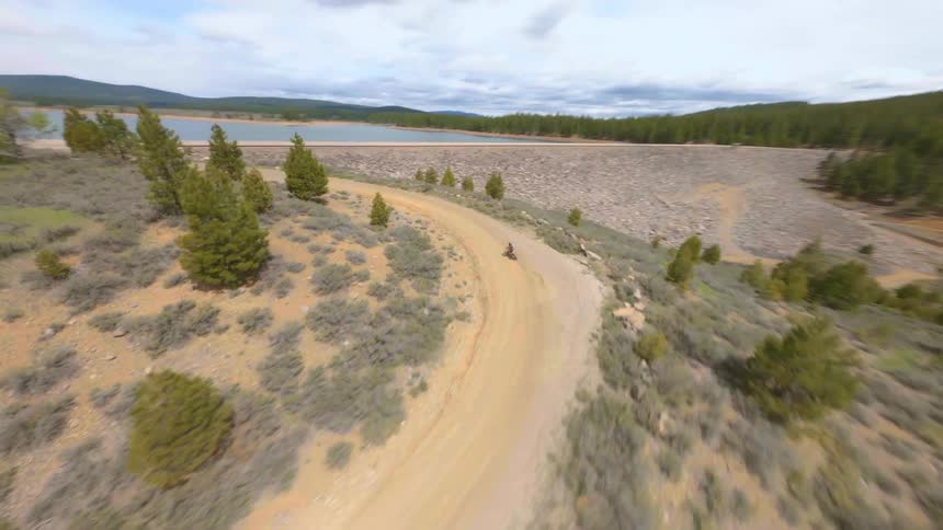 Man drives a motorcycle at full speed on a dirt road near a dam
