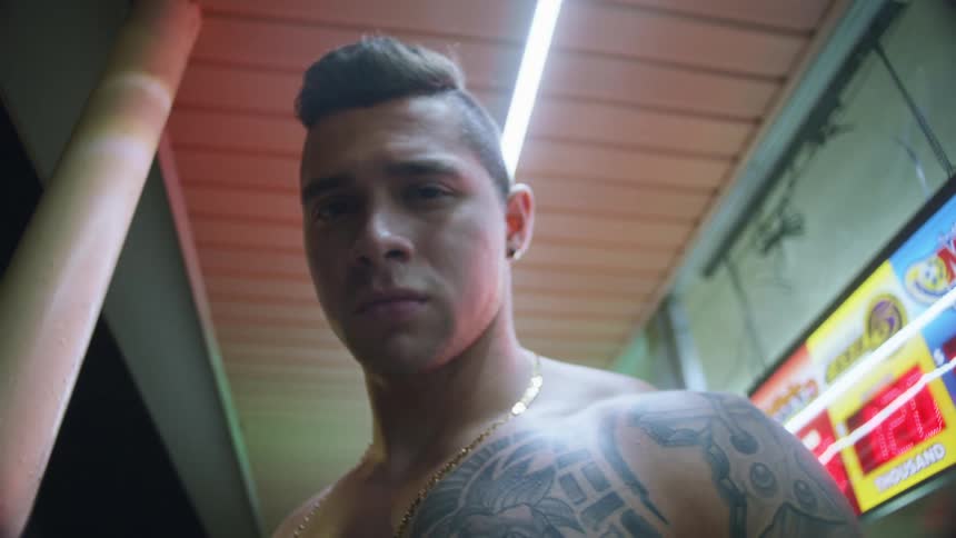 Shirtless man with tattoos and gold chain looking down at camera outside convenience store at night