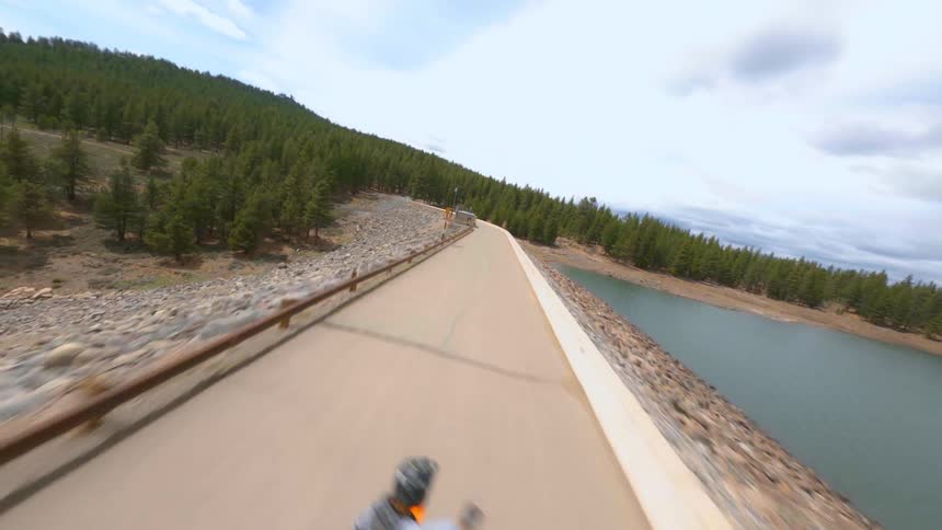 A person standing while riding a speeding dirt bike down a narrow road next to a lake waves at the drone filming them as it passes by