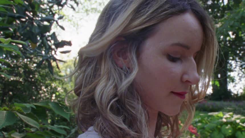 Blonde woman with a curly hair looks around and smiles slightly in the public park