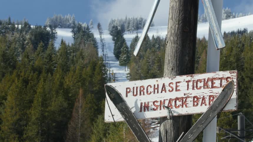 Ski resort sign on a pole near a sky lift going up the mountains between pine tree forests