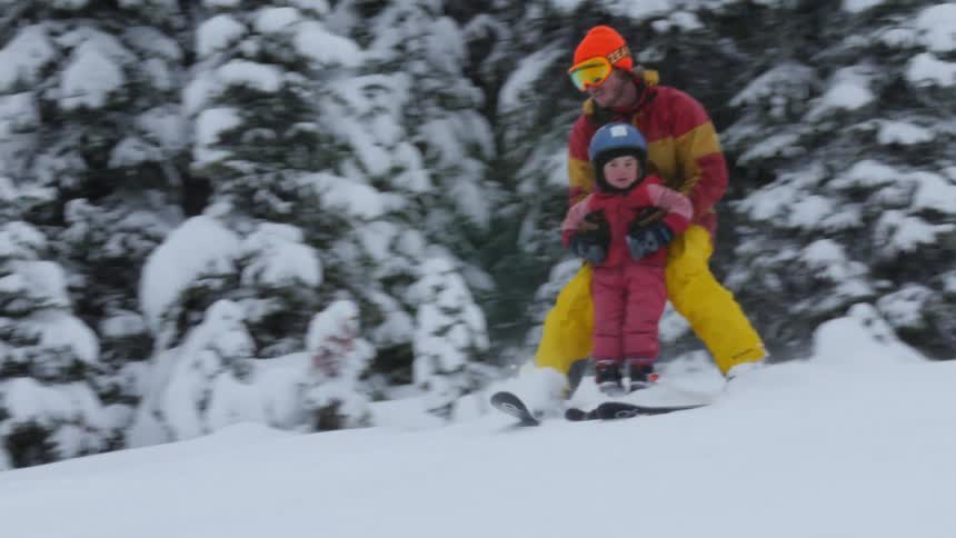 A man skis down a hill while helping his daughter while his other daughter skis on her own