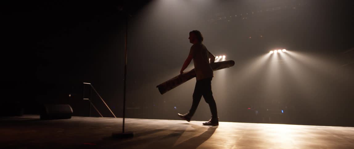 A stagehand carries a rolled up rug across the stage of and auditorium
