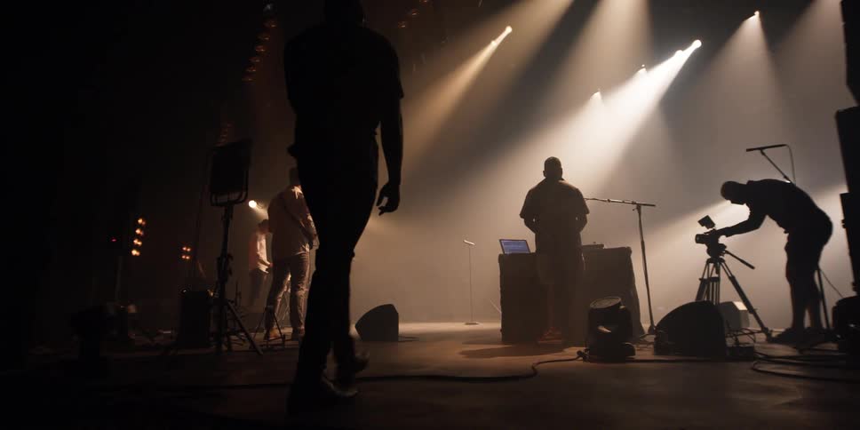 Men stand on shadowy stage adjusting musical instruments under spotlights