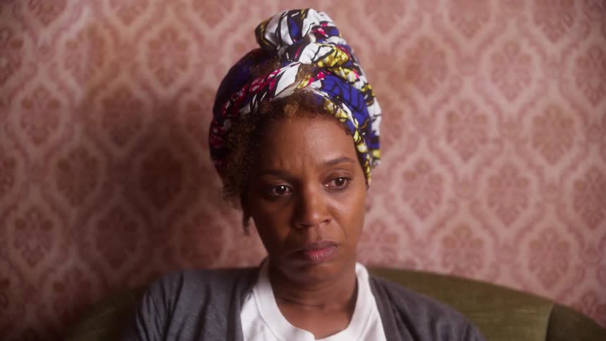 A black woman in a headwrap shakes her head while brooding in a chair