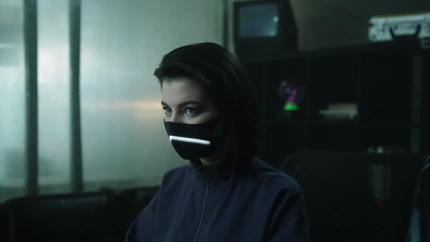 Woman wearing a face mask sits in a control room while white lights scan her face