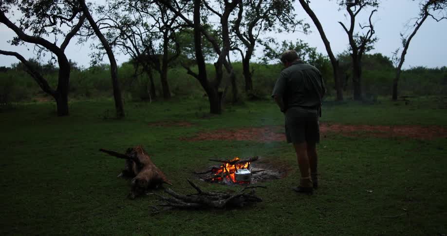 Man stands and waits by the a campfire to cook food with a pot